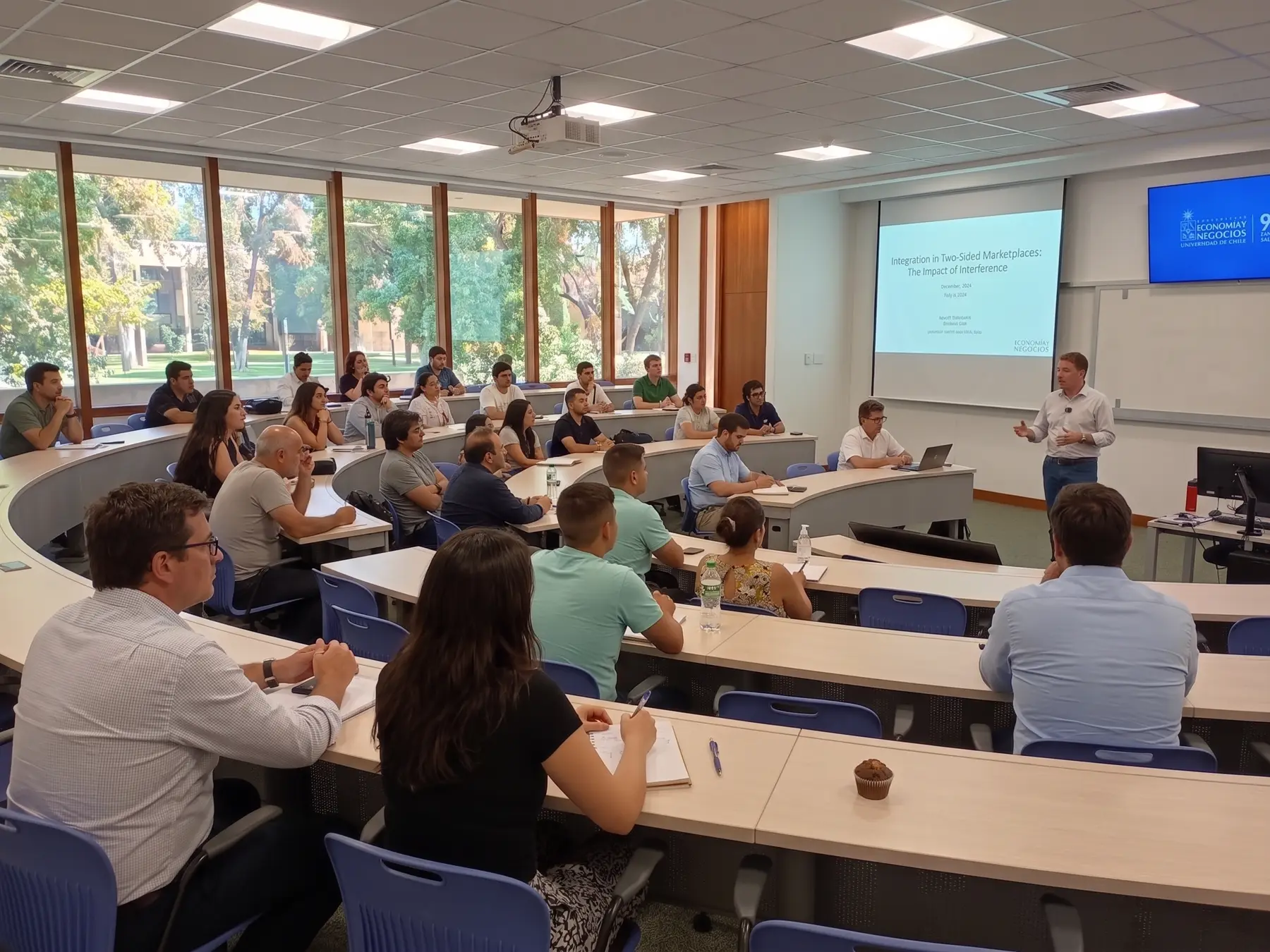 Sala de clases con ventanales, gente sentada siguiendo una presentación.